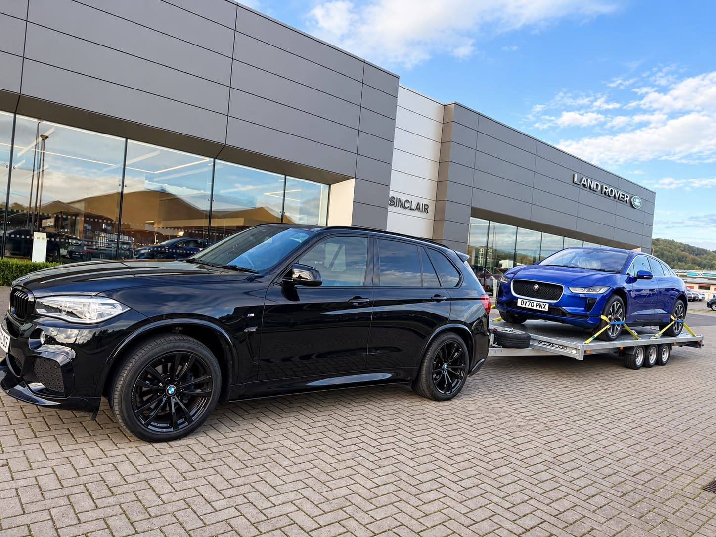 A black BMW X5 towing a blue Jaguar I-Pace outside a Land Rover dealership